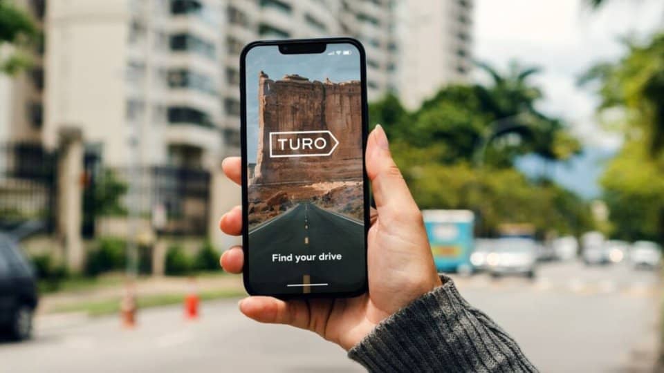 image showing a woman's hand holding a phone with the Turo logo on it - header graphic for how to make money on Turo post on ridester.com